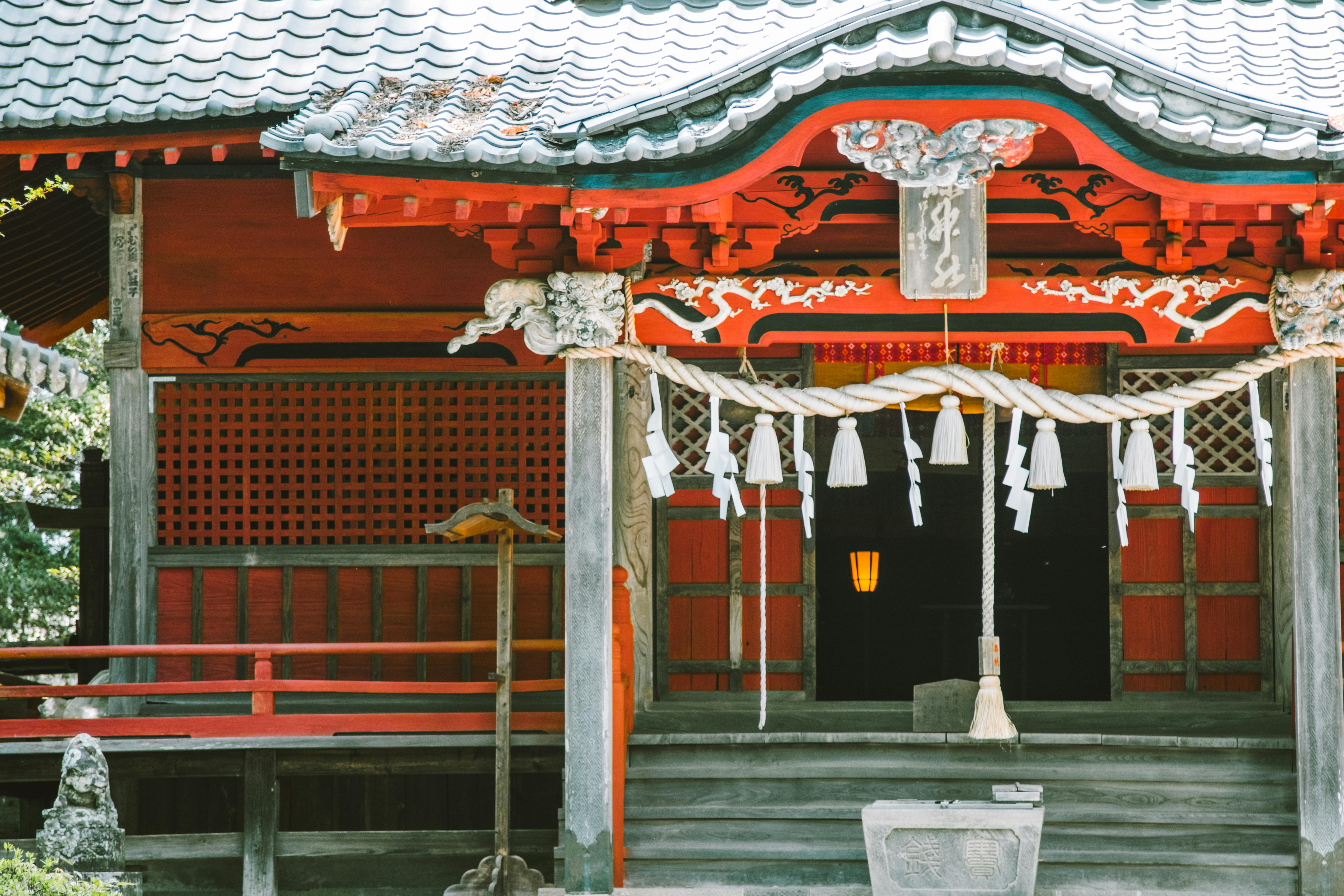 A traditional Japanese Shinto shrine with red wooden walls, ornate carvings, a tiled roof, and decorative white paper streamers hanging on a rope above the entrance.