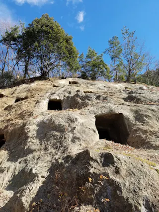 Rocky hillside with several rectangular cave openings carved into the surface. Sparse trees grow at the top and blue sky with a few clouds is visible above. The scene suggests an ancient or historical site.