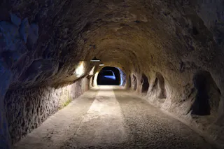 A dimly lit tunnel carved from rock, with rough stone walls and ceiling, illuminated by a few overhead lamps, leading to a metal gate at the far end.
