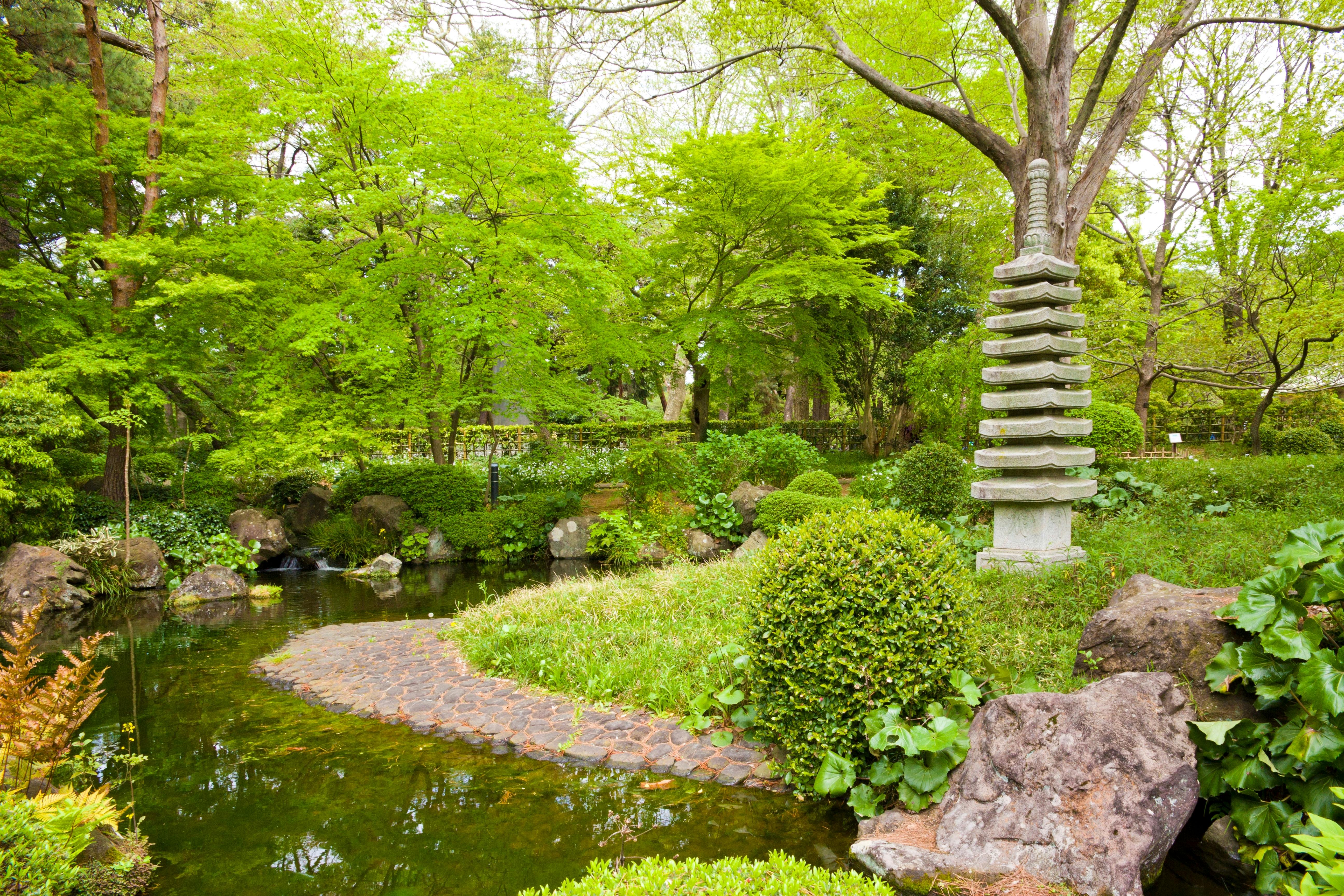 A lush Japanese garden with vibrant green trees, a stone path curving over a pond, manicured bushes, rocks, and a tall stone pagoda sculpture on the right.
