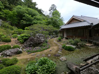 A traditional Japanese house stands beside a lush garden with green shrubs, trees, rocks, and a winding stone path under a cloudy sky.