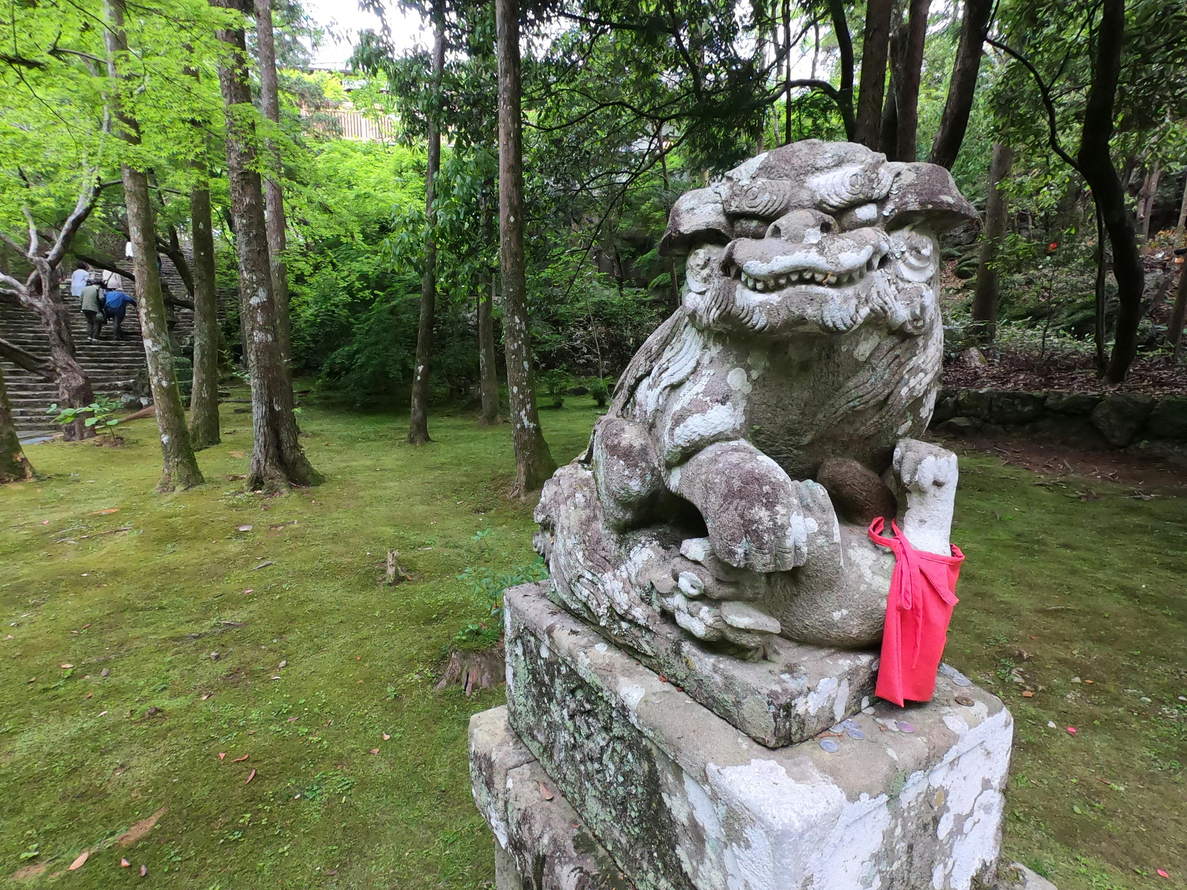A stone guardian lion statue with a red cloth tied around its leg sits on a pedestal in a lush, green forest. Stone steps and people are visible in the background among the trees.