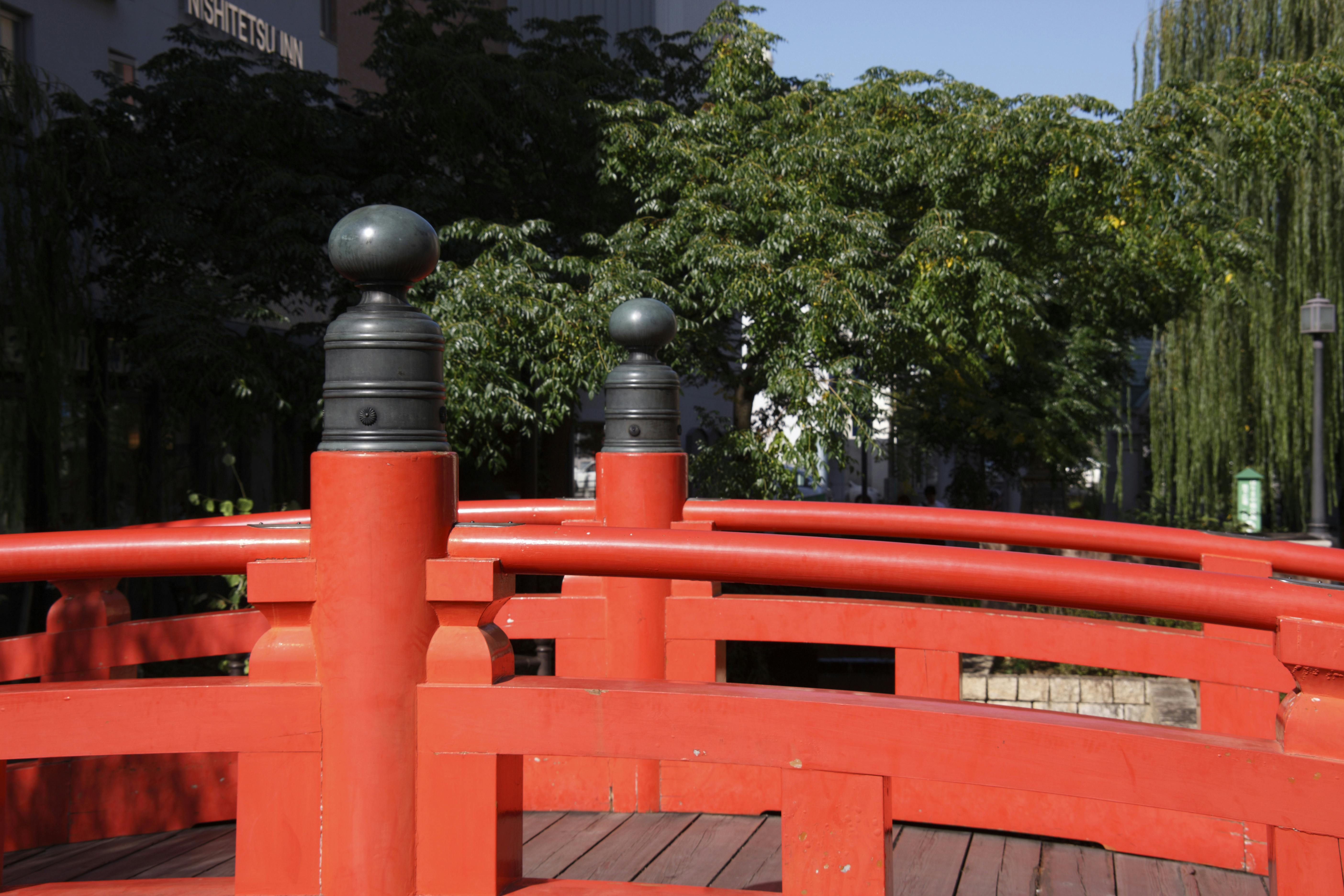 A bright red arched wooden bridge with black finials spans over a walkway, surrounded by lush green trees under a clear blue sky.