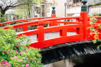 A vibrant red wooden bridge arches over a narrow stream, surrounded by green foliage and blooming flowers, with nearby buildings in the background under a bright, clear sky.