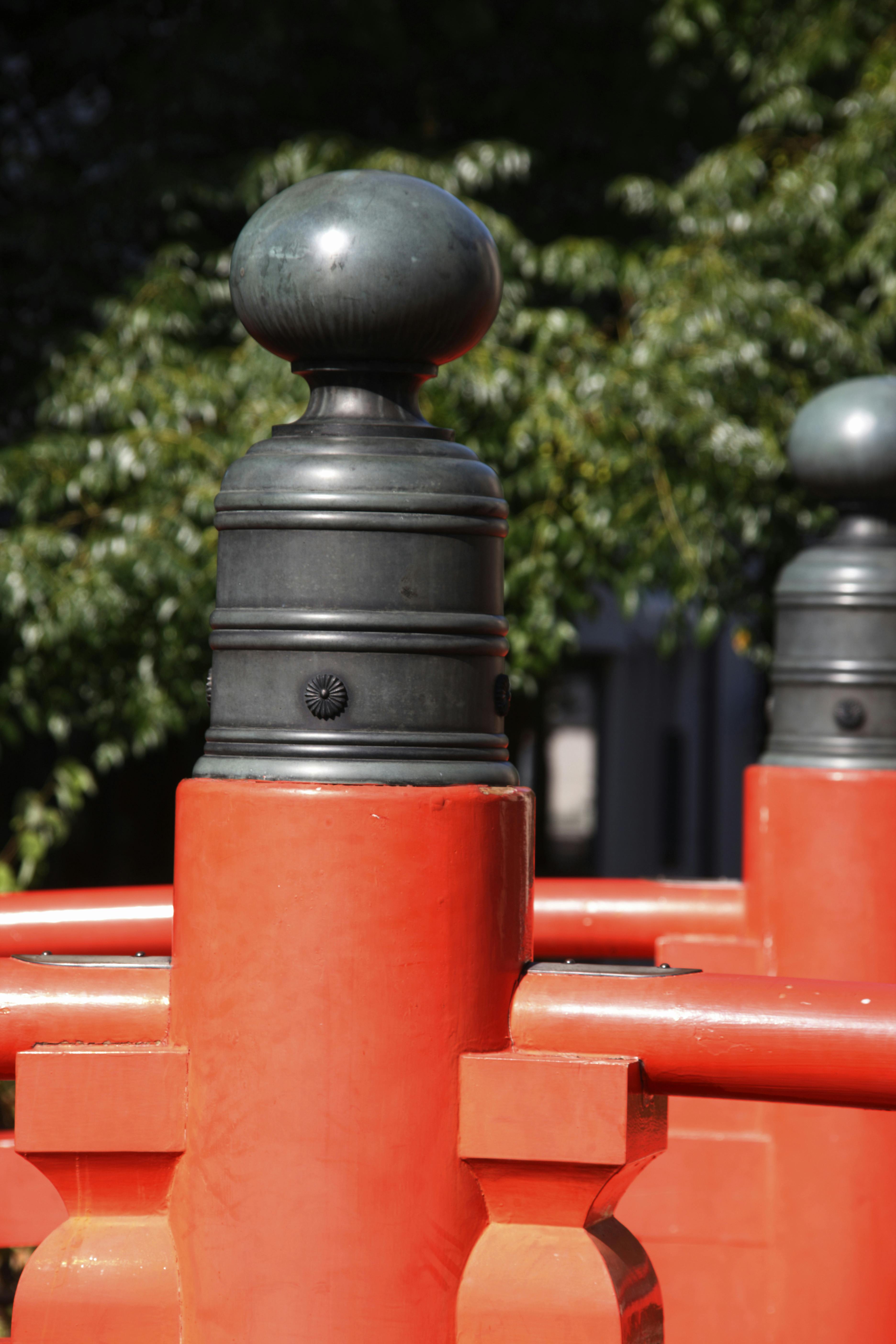 A close-up view of a decorative black metal post atop a bright red-painted railing, with green trees blurred in the background. The design suggests traditional Japanese architecture.