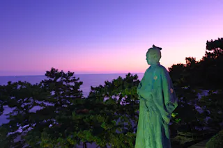 A bronze statue of a man in traditional Japanese clothing overlooks the ocean at sunset, surrounded by pine trees and under a purple and pink sky.