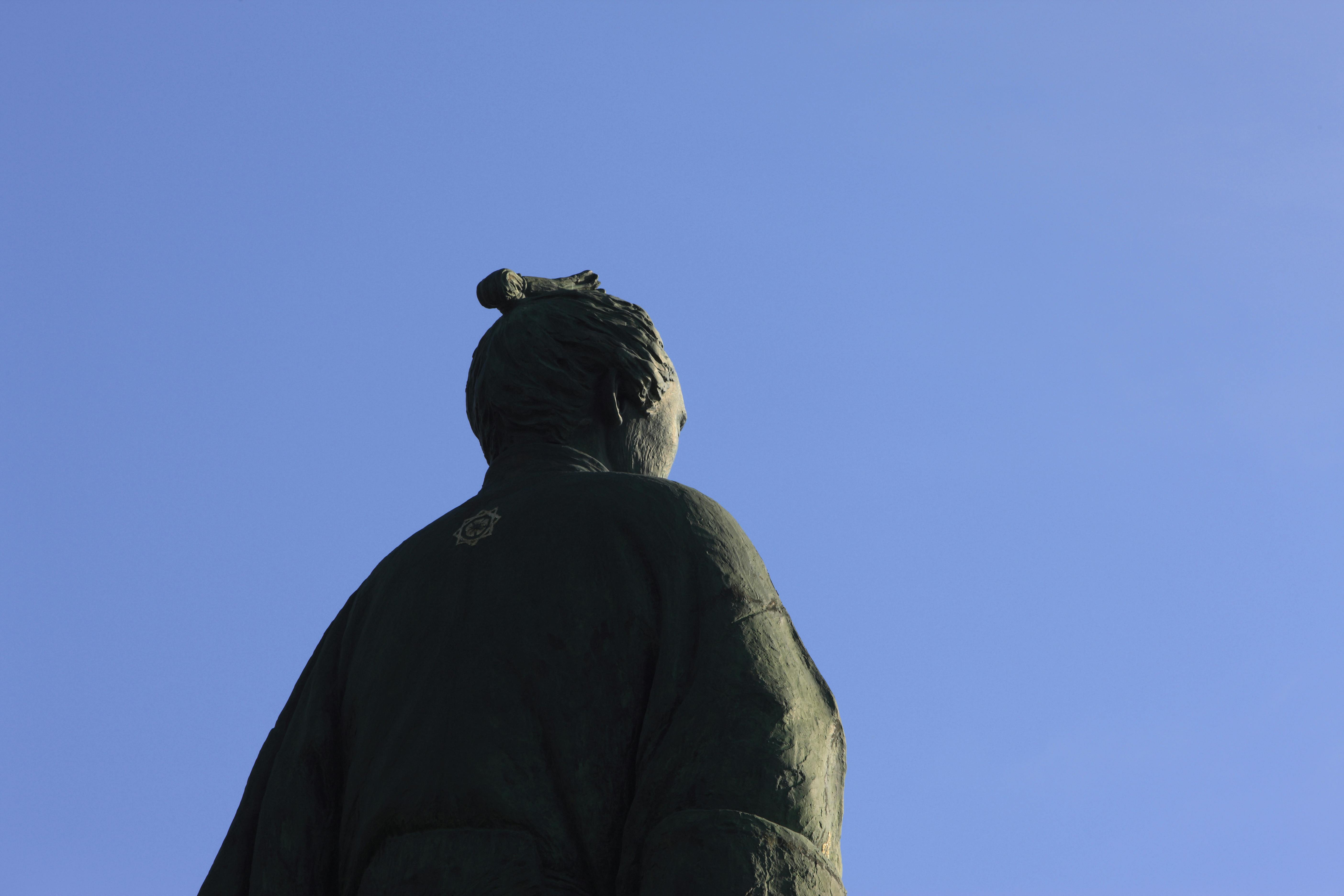 A bronze statue of a person in traditional clothing is seen from behind against a clear blue sky. The figure’s hair is styled in a traditional topknot, and a small emblem is visible on the upper back of the garment.