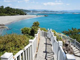 A concrete staircase with white railings leads down to a sandy beach and turquoise sea, surrounded by green foliage, on a sunny day with mountains visible in the background.