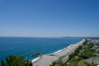 A wide view of a coastline with clear blue water, sandy beaches, and a road running alongside. Trees and greenery border the road, and the sky is clear and blue. Concrete barriers extend into the sea.