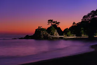 A rocky coastline at sunset with trees atop the rocks, a path winding around them, and the sky glowing in shades of orange, pink, and purple, reflecting on the calm water below.