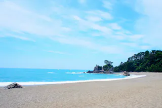 A sandy beach with gentle waves, clear blue water, and a partly cloudy sky. There are rocks and trees along the shoreline in the distance.