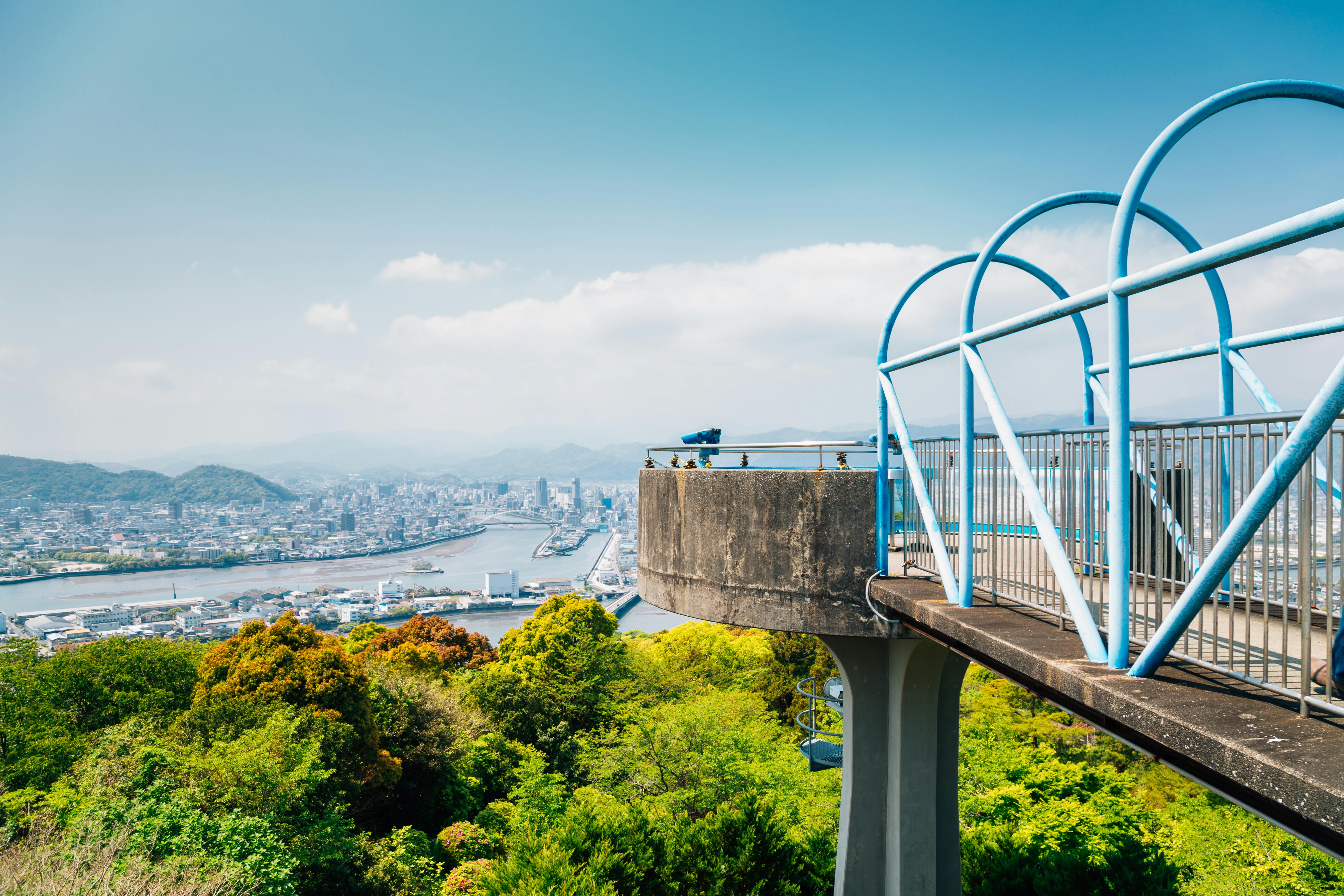 A blue observation deck extends over lush green trees, offering a panoramic view of a city, river, and distant mountains under a clear, blue sky.