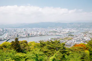 A panoramic view of a city with rivers running through it, surrounded by lush green trees in the foreground and mountains in the background under a partly cloudy sky.