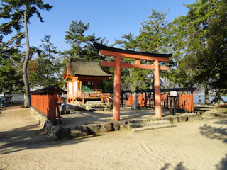 A traditional Japanese torii gate stands before a small shrine with a thatched roof, surrounded by a wooden fence and pine trees under a clear blue sky.