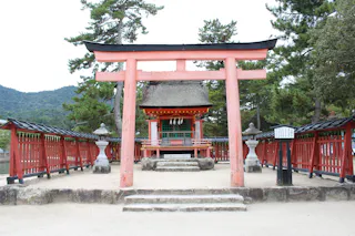 A traditional Japanese Shinto shrine with a red torii gate, stone steps, and red fences, surrounded by trees and mountains in the background.