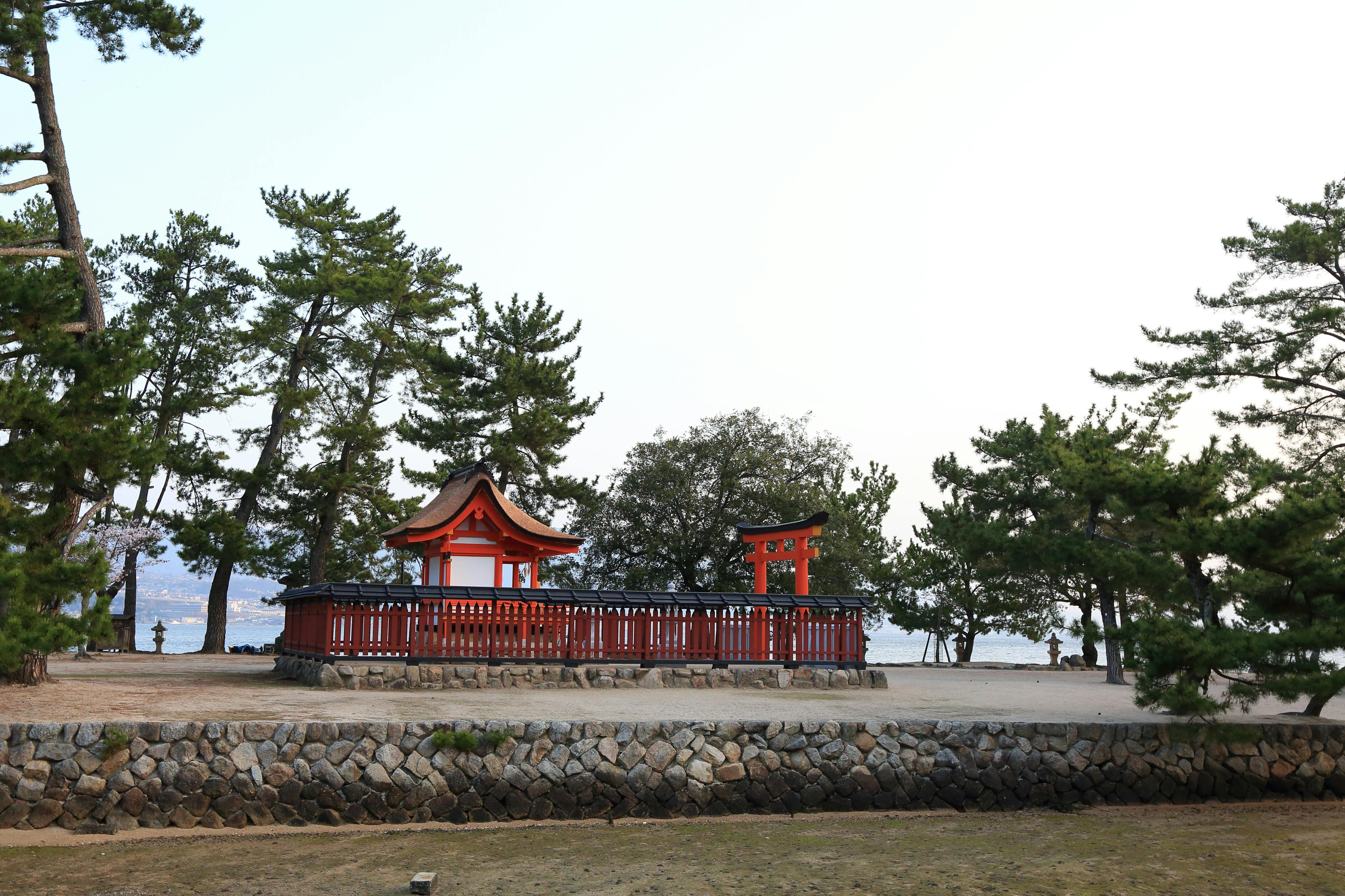 A small traditional Japanese shrine with a red torii gate is surrounded by trees and stone walls, set on a raised area near the water under a pale sky.
