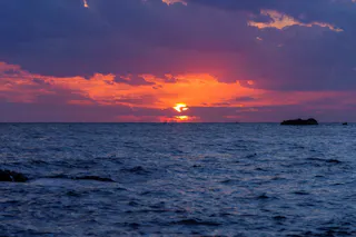 The sun sets over a calm ocean, casting vibrant orange and purple hues across the sky and water, with dark clouds and distant rocks on the horizon.