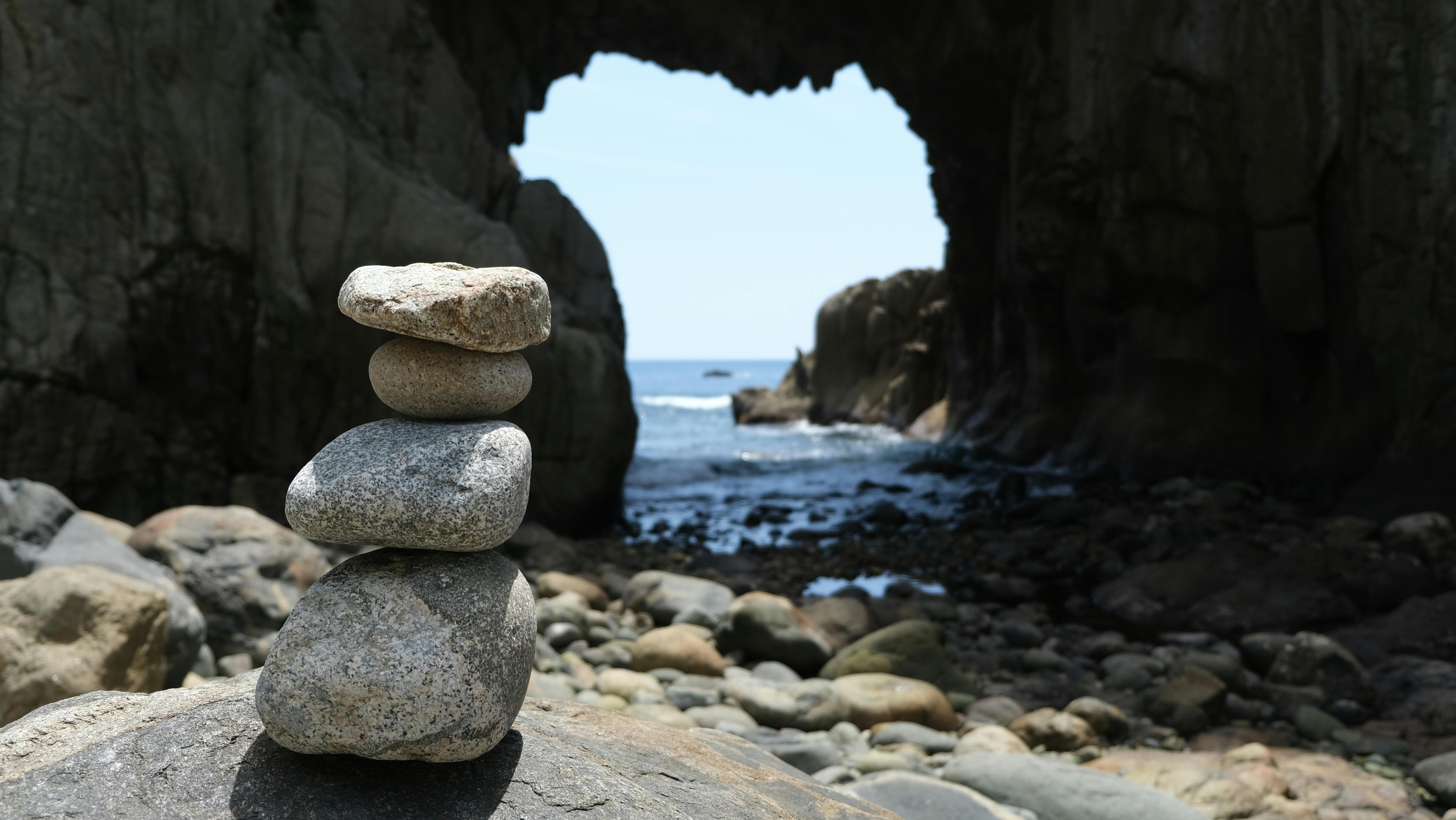A stack of four smooth stones is balanced on a large rock in the foreground, with a rocky archway and the ocean visible in the background under daylight.