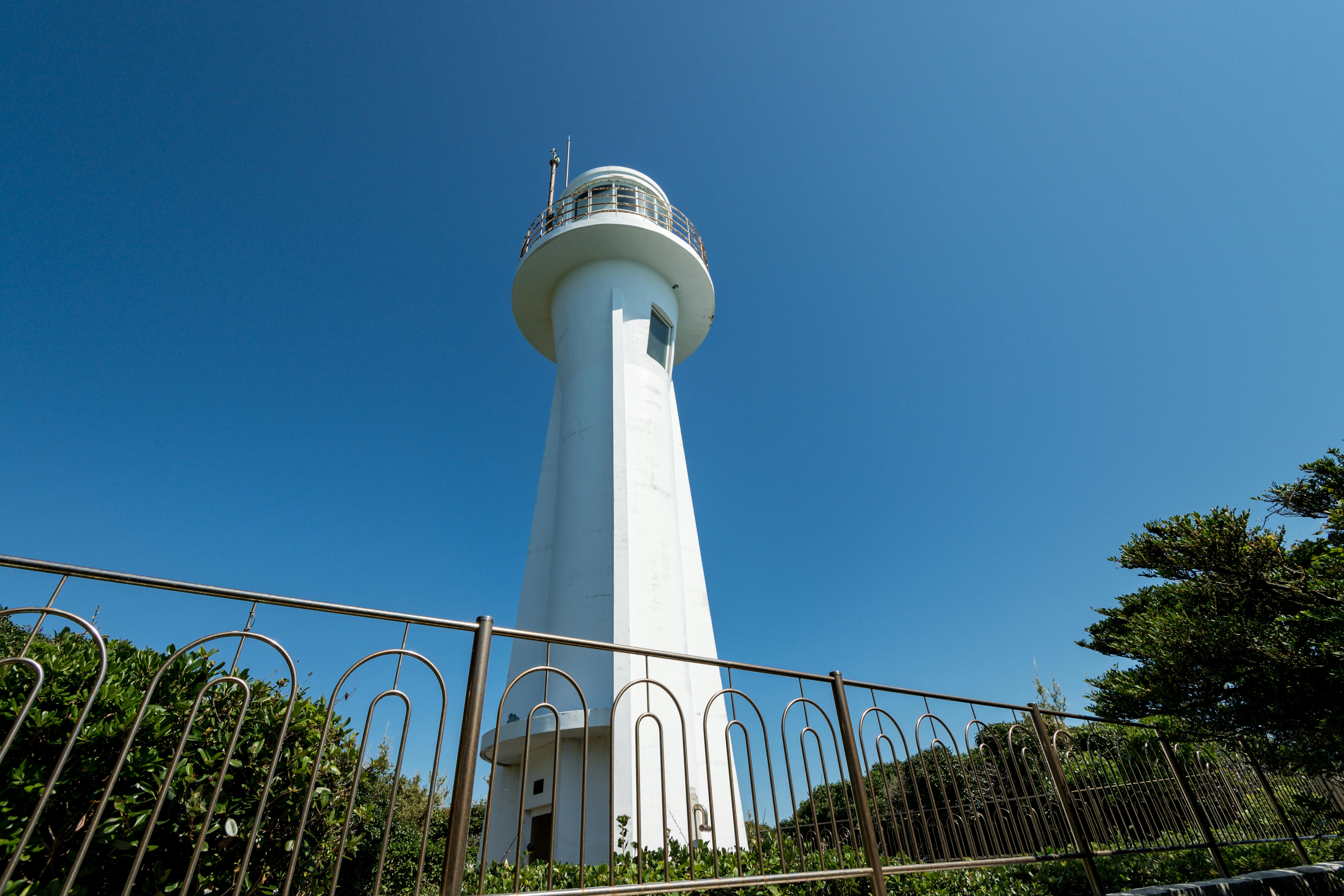 A tall white lighthouse stands against a clear blue sky, surrounded by greenery and enclosed by a metal fence in the foreground.