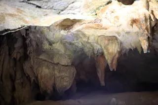 Jagged stalactites and rock formations hang from the ceiling of a dimly lit cave, with rough, textured surfaces in earthy tones of brown and tan.