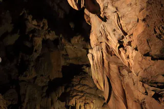 Rocky cave interior with rough, textured brown walls and shadows, illuminated by soft light highlighting natural formations and crevices.