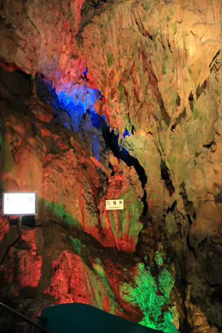 A cave interior with jagged rock formations illuminated by colorful lights in red, green, and blue. Two signs with Japanese writing are visible among the rocks.