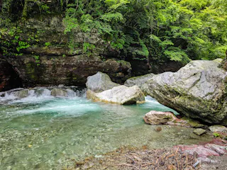 Clear turquoise water flows gently over rocks in a forested area, surrounded by large boulders and lush green vegetation on a sunny day. The scene is peaceful and natural.