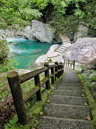 A mossy stone staircase with a wooden handrail leads down to a turquoise river surrounded by rocks and lush green trees. A small stone bridge crosses the water in the background.