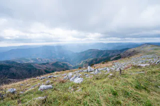 A grassy mountain slope with scattered rocks overlooks a vast valley of rolling hills and distant mountains under a cloudy, overcast sky.