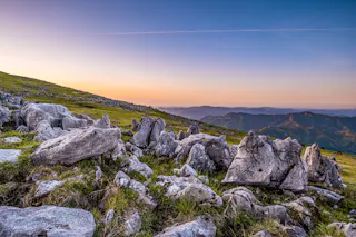 Rocky landscape with scattered boulders and grass in the foreground, gentle hills and mountains fading into the distance, and a colorful gradient sunset sky overhead with a thin streak of cloud.