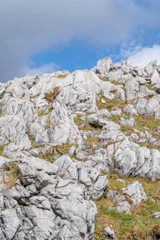 Rocky hillside with jagged white stones and patches of green grass under a partly cloudy blue sky.