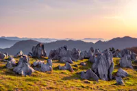 Jagged gray rock formations scattered across a grassy hilltop at sunrise, with layers of rolling, tree-covered mountains fading into the misty distance under a colorful sky.