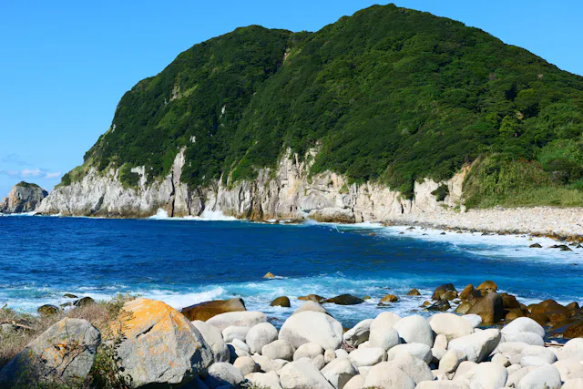 A rocky shoreline with large white boulders, blue ocean waves, and a steep, green-covered hill rising in the background under a clear blue sky.