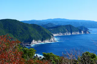 A scenic coastline with lush green hills meeting the deep blue sea, waves crashing against rocky cliffs, and mountains visible in the background under a clear sky. Some red and green foliage appears in the foreground.