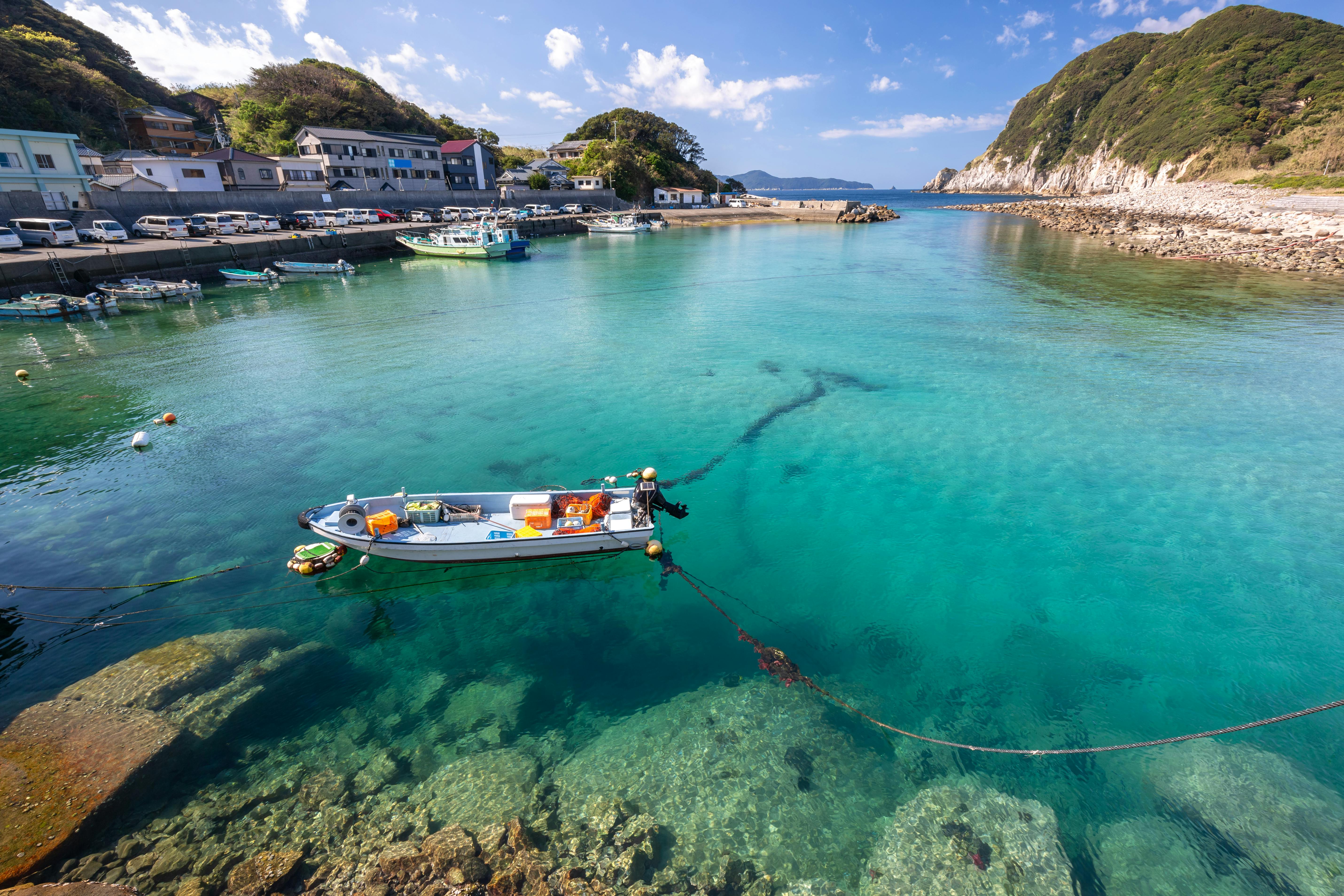 A small fishing boat floats on clear turquoise water in a harbor, with rocky shores, parked cars, and houses in the background, surrounded by green hills and a partly cloudy sky.