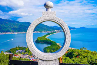A stone circular monument frames a view of a scenic sandbar lined with trees stretching into blue water, surrounded by green mountains under a partly cloudy sky.