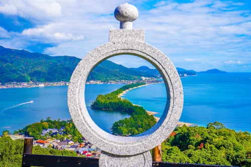 A stone circular monument frames a view of a scenic sandbar lined with trees stretching into blue water, surrounded by green mountains under a partly cloudy sky.