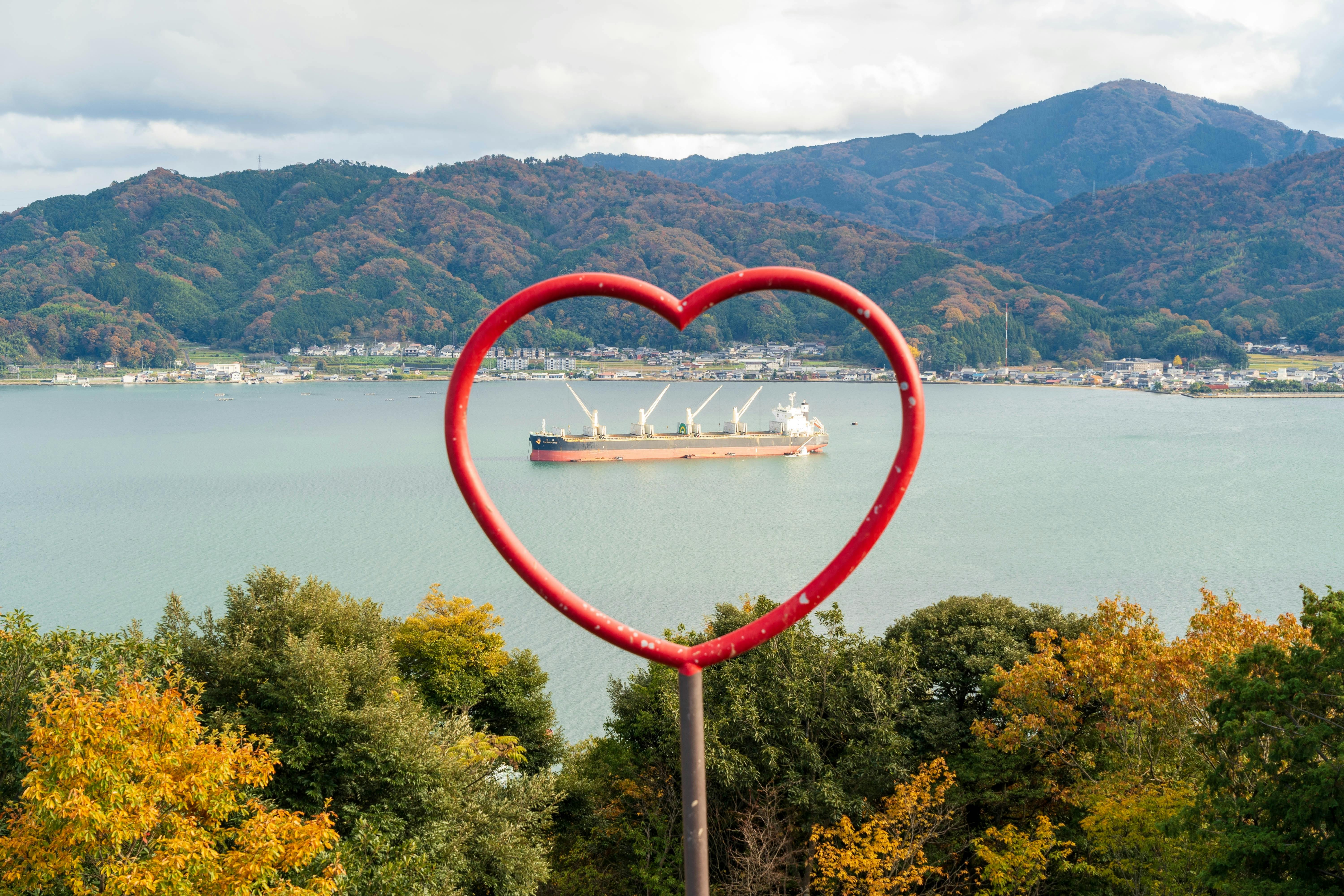 A large red heart-shaped frame stands in the foreground, overlooking a body of water with a cargo ship and forested hills in the background during autumn.