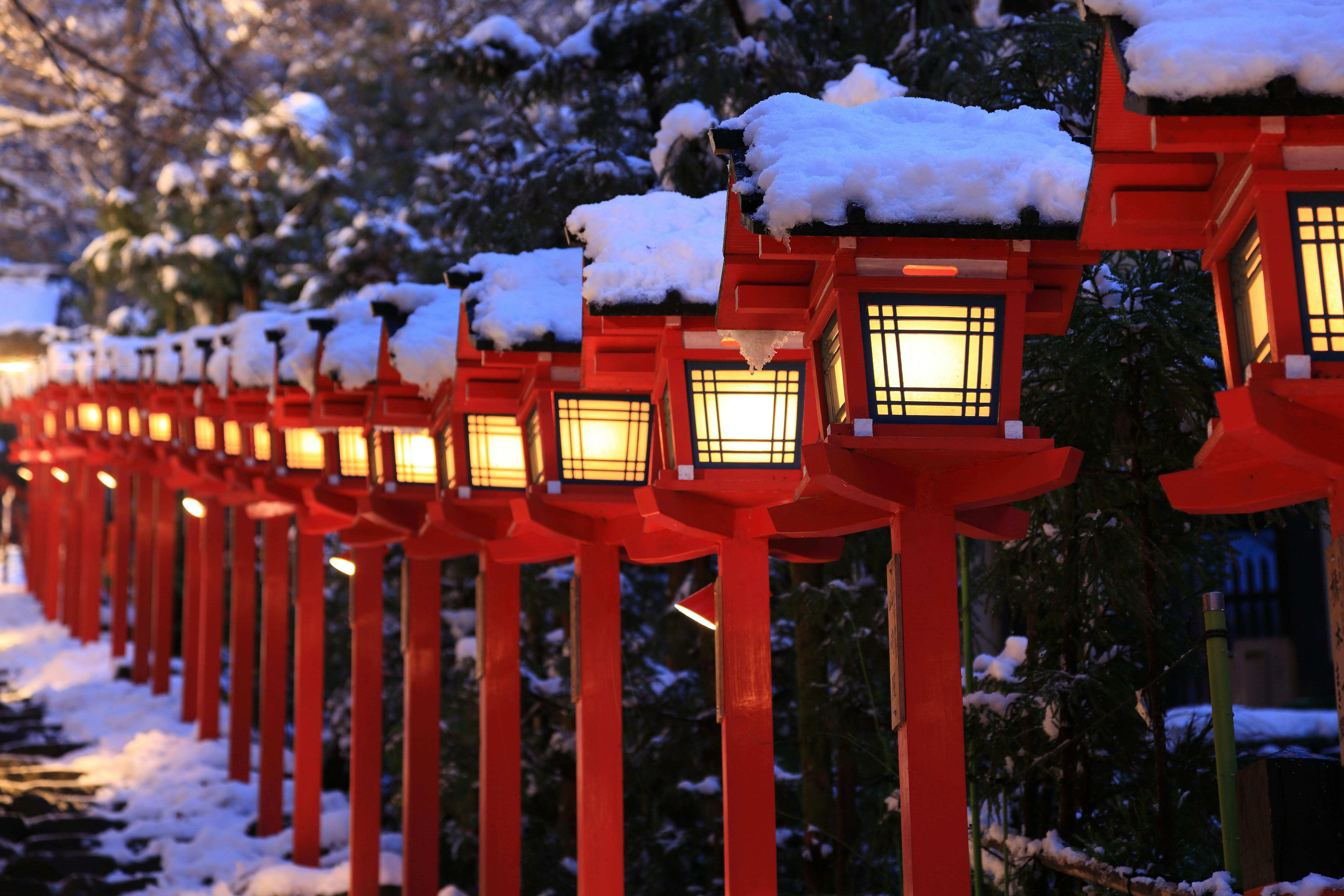 A row of traditional Japanese lanterns, painted red and topped with snow, lines a stone path at dusk, casting a warm glow amid surrounding trees and winter scenery.