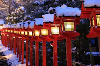 A row of traditional Japanese lanterns, painted red and topped with snow, lines a stone path at dusk, casting a warm glow amid surrounding trees and winter scenery.