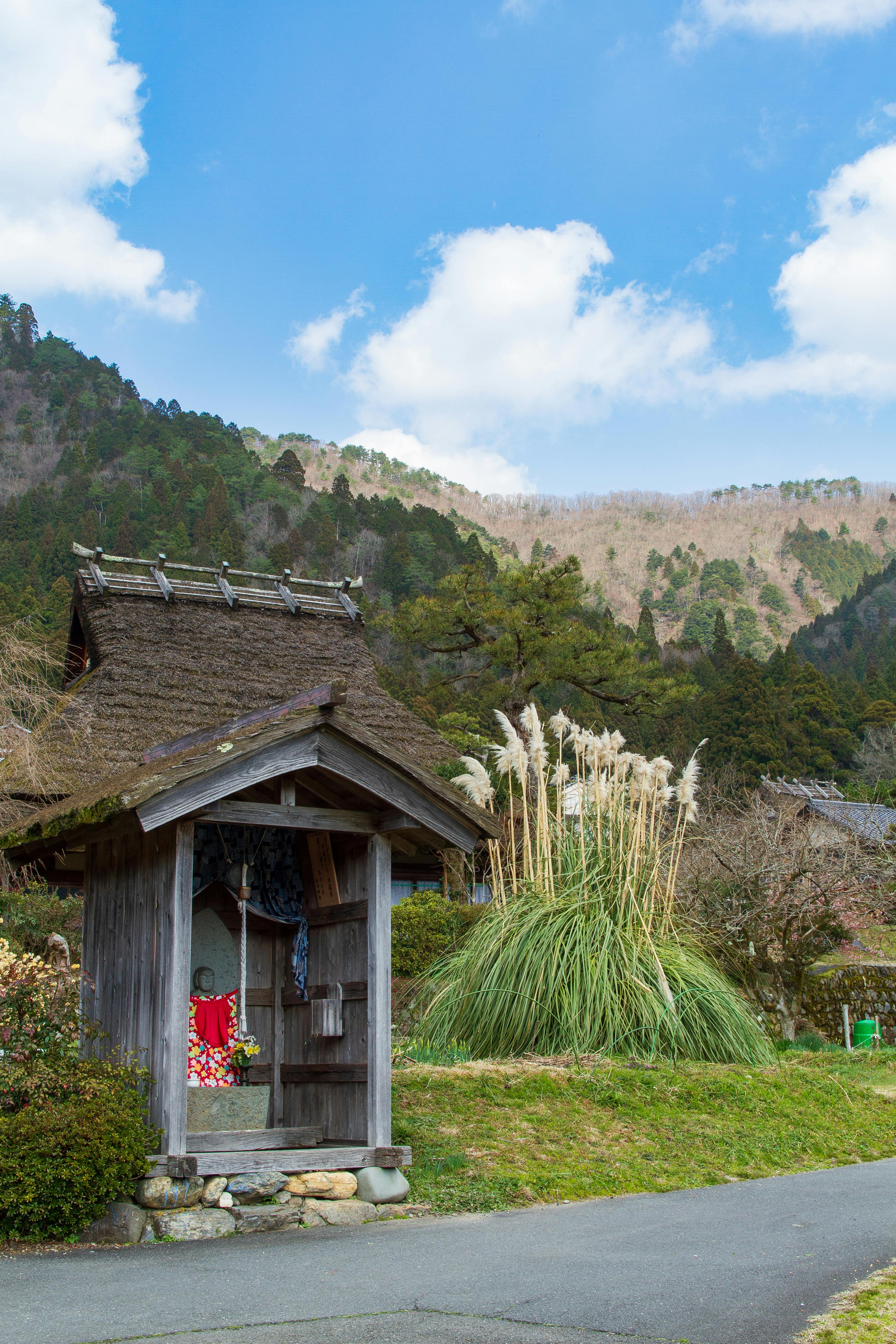 A small wooden shrine with a thatched roof stands by a rural roadside, surrounded by tall grasses and greenery, with forested mountains and a blue sky with scattered clouds in the background.