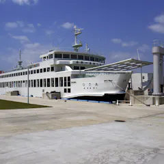 Port Miyanoura A large white ferry docked at a concrete pier under a partly cloudy sky, with a gangway extended for boarding and green grass visible on the left side.