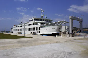 Port Miyanoura A large white ferry docked at a concrete pier under a partly cloudy sky, with a gangway extended for boarding and green grass visible on the left side.