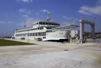 A large white ferry docked at a concrete pier under a partly cloudy sky, with a gangway extended for boarding and green grass visible on the left side.