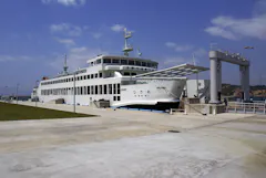 A large white ferry docked at a concrete pier under a partly cloudy sky, with a gangway extended for boarding and green grass visible on the left side.