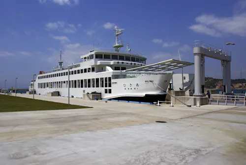A large white ferry docked at a concrete pier under a partly cloudy sky, with a gangway extended for boarding and green grass visible on the left side.