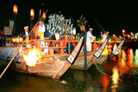 A traditional wooden boat decorated with lanterns and ornaments carries people in traditional Japanese attire, with a large fire at the bow, floating on water at night during a festival.