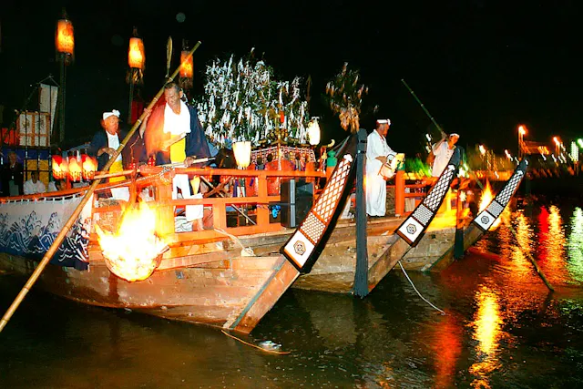 A traditional wooden boat decorated with lanterns and ornaments carries people in traditional Japanese attire, with a large fire at the bow, floating on water at night during a festival.