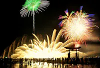 A crowd watches vibrant fireworks illuminating the night sky above a traditional Japanese torii gate, with bright green, gold, and multicolored bursts reflecting on water.