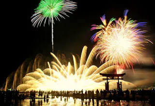 A crowd watches vibrant fireworks illuminating the night sky above a traditional Japanese torii gate, with bright green, gold, and multicolored bursts reflecting on water.